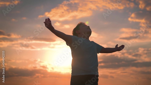 Child raised his hands to sky in park at sunset, true faith. Child plays in park against sky. Little boy prays against background of sky and sun. Religion and god, childhood dreams. Happy family