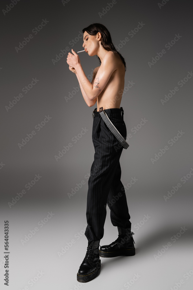 full length of shirtless woman in black trousers and rough boots lighting cigarette on grey background. full length of shirtless woman in black trousers and rough boots lighting cigarette on grey background.