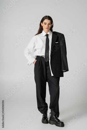 full length of young model in oversize formal wear and rough boots standing with hand in pocket on grey background.
