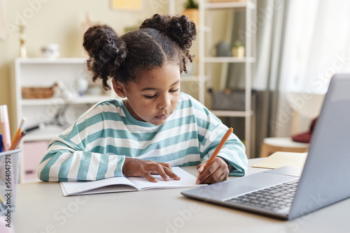 Cute black girl writing in notebook doing homework at desk in home interior