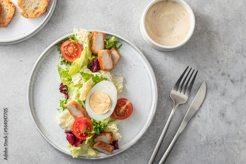 Caesar salad in modern linear feed with chicken, egg, tomatoes, lettuce and parmesan chips with cutlery on gray table, top view, flat lay, menu