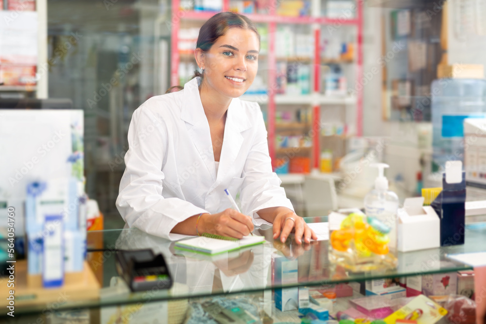 Female pharmacist in gown standing behind counter in drugstore. She's writing recipe.