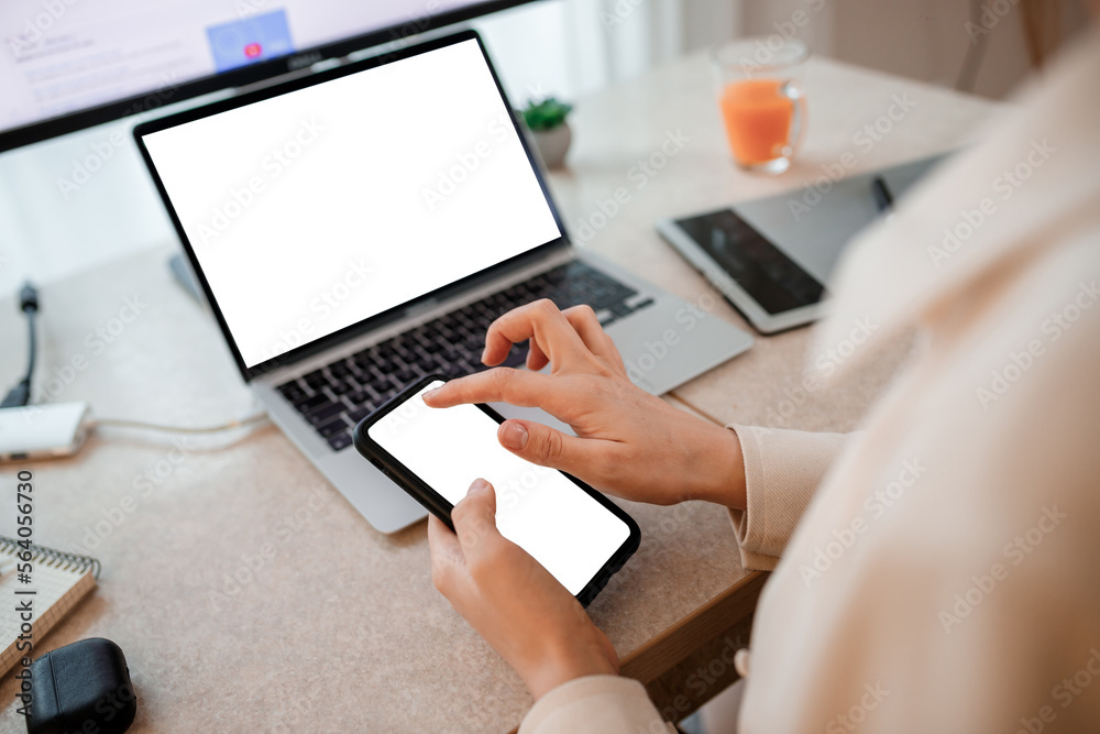 Close up of businesswoman working at home office on laptop and making notes on phone. Young happy female student studying in online university