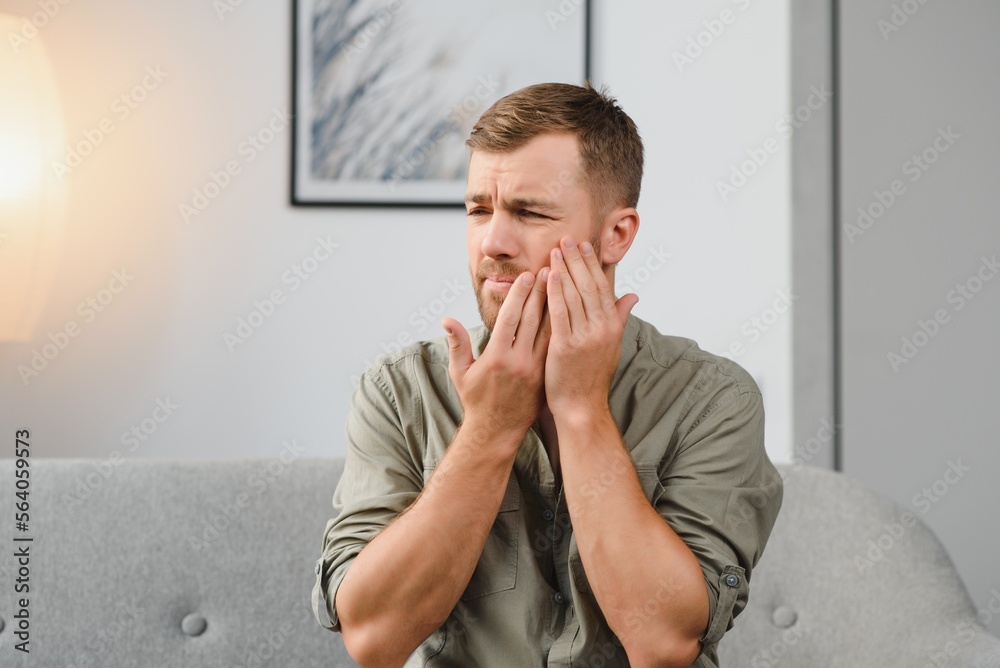 Dental health problems concept. Man suffering from strong toothache. Upset male patient with inflamed nerve, tooth abscess, or teeth sensitivity sitting on sofa and touching cheek with grimace of pain Dental health problems concept. Man suffering from strong toothache. Upset male patient with inflamed nerve, tooth abscess, or teeth sensitivity sitting on sofa and touching cheek with grimace of pain
