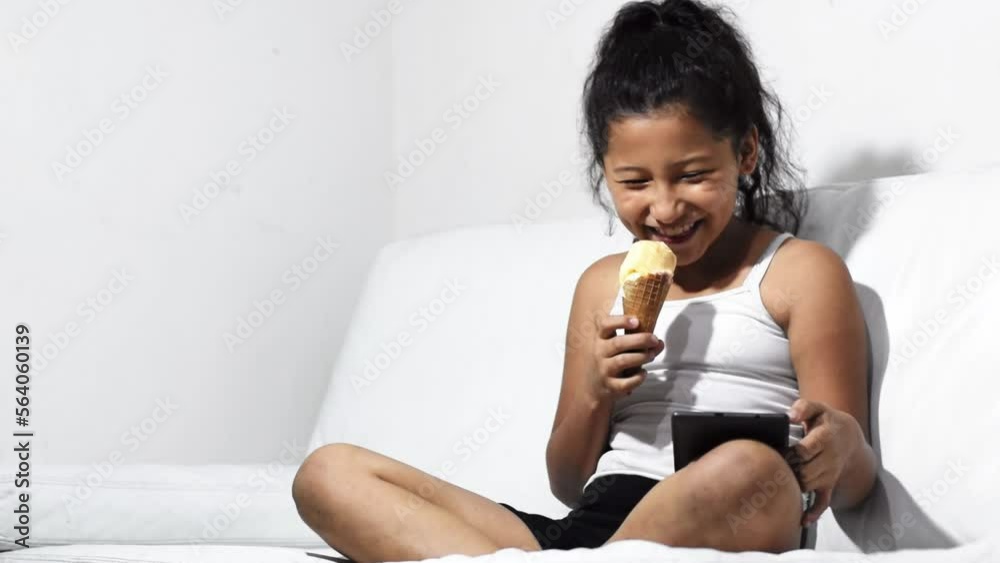 little latina girl, brunette with an impressed face opening her mouth in excitement while eating her ice cream and looking at her tablet sitting on the sofa at home. little latina girl, brunette with an impressed face opening her mouth in excitement while eating her ice cream and looking at her tablet sitting on the sofa at home.