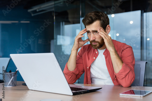 Frustrated businessman depressed at workplace working on laptop, man in shirt upset and sad displeased with bad work results and achievement inside office.