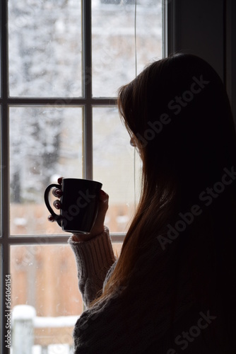 Young woman looking out of the window on a snow day. Pensive lady drinking coffee while gazing outside