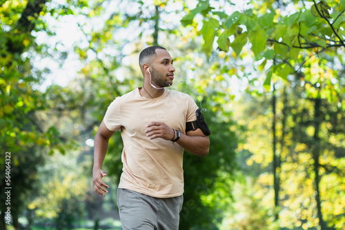 Hispanic man jogging in the park on a sunny day, runner listening to music in wired headphones, audio books and podcasts, sportsman happy with an active weekend.