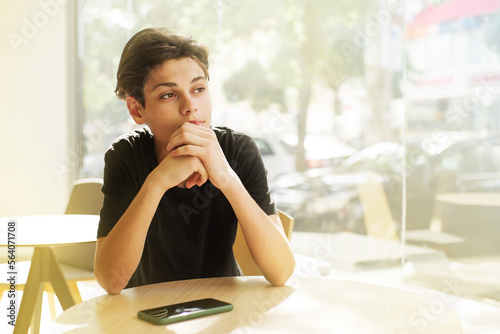 Young handsome guy, a teenager, sits at a table in a cafe and looks thoughtfully out the window on a sunny day