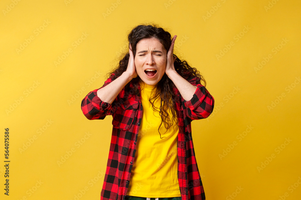 young curly girl covers her ears with her hands and screams on a yellow background, student girl in stress is hysterical