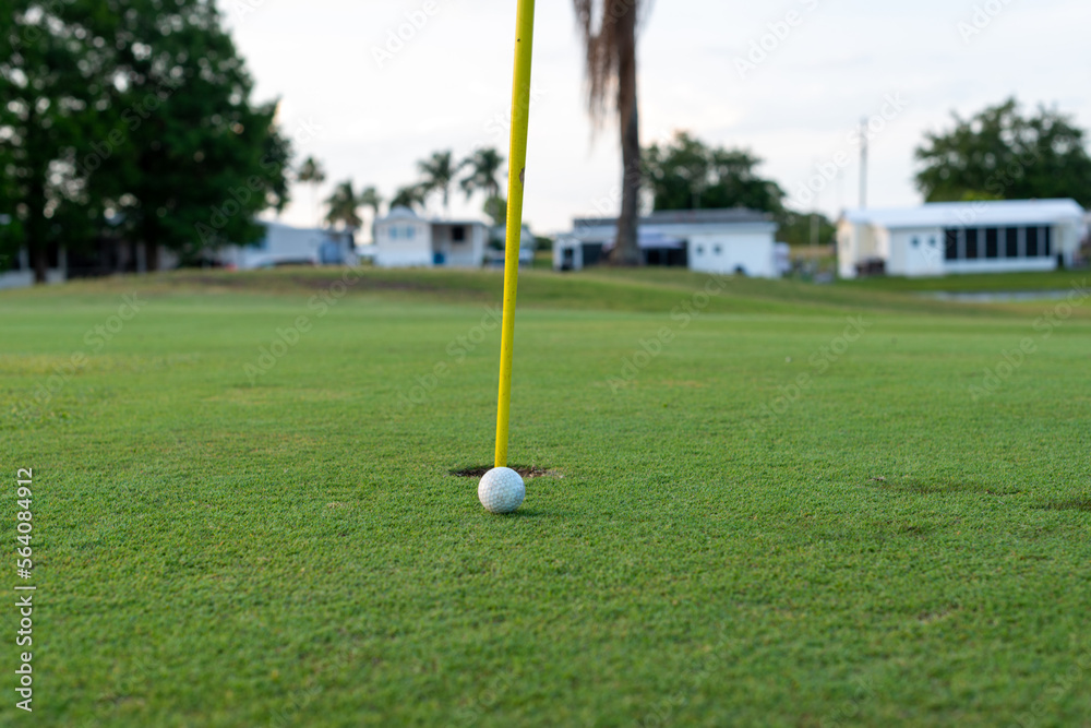 Obraz premium A small white golf ball sits on lush green golf grass on the finishing hole. The ball is close to the hole with a yellow metal flag pole in the hole. The thin pole is tilted in the circular cup.