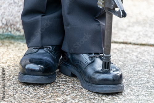 A military person stands at attention with his rifle pitched on the toe of his shinned boot. The male navy officer stands on guard wearing dark pants, black boots, and a rifle resting on the uniform.