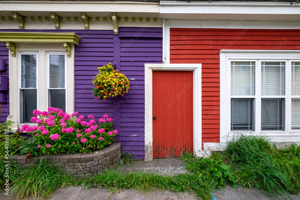 A purple exterior clapboard wooden wall with a hanging flower basket. There's a red door joining the purple wall to a red clapboard wall with a double hung window. There are a flower box and grass too A purple exterior clapboard wooden wall with a hanging flower basket. There's a red door joining the purple wall to a red clapboard wall with a double hung window. There are a flower box and grass too