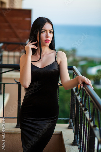 Beautiful sexy young long-haired brunette caucasian girl in slimming black dress attractively posing at Antalya hotel's balcony with softly blurred blueish sea background