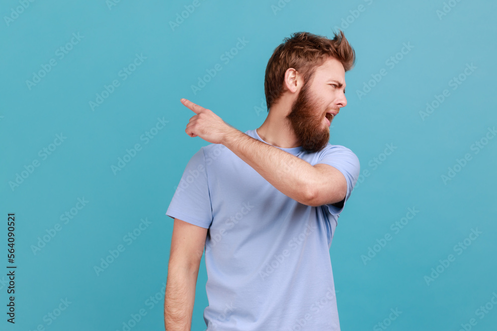Get out. Portrait of handsome bearded man looking aside and pointing finger another way, asking to leave, dissatisfied expression. Indoor studio shot isolated on blue background. Get out. Portrait of handsome bearded man looking aside and pointing finger another way, asking to leave, dissatisfied expression. Indoor studio shot isolated on blue background.