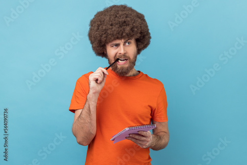 Portrait of man with Afro hairstyle in orange T-shirt standing thoughtful biting pencil, pondering idea to write into notebook, taking notes in paper. Indoor studio shot isolated on blue background. photo