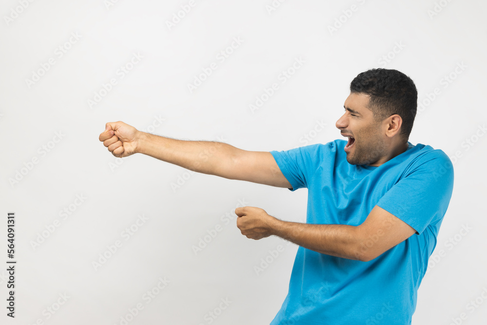 Side view portrait of strong attractive young adult unshaven man wearing blue T- shirt standing pulling, trying to pull something heavy. Indoor studio shot isolated on gray background. Side view portrait of strong attractive young adult unshaven man wearing blue T- shirt standing pulling, trying to pull something heavy. Indoor studio shot isolated on gray background.