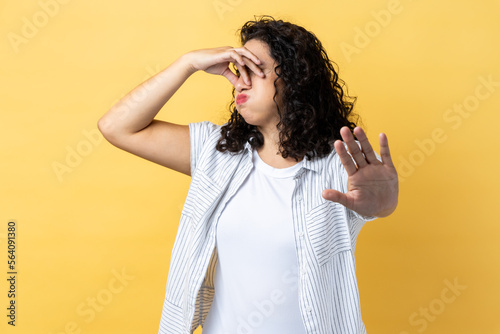 Portrait of woman with dark wavy hair pinching her nose to hold breath and showing stop gesture, disgusted by unpleasant odor, fart. Indoor studio shot isolated on yellow background.