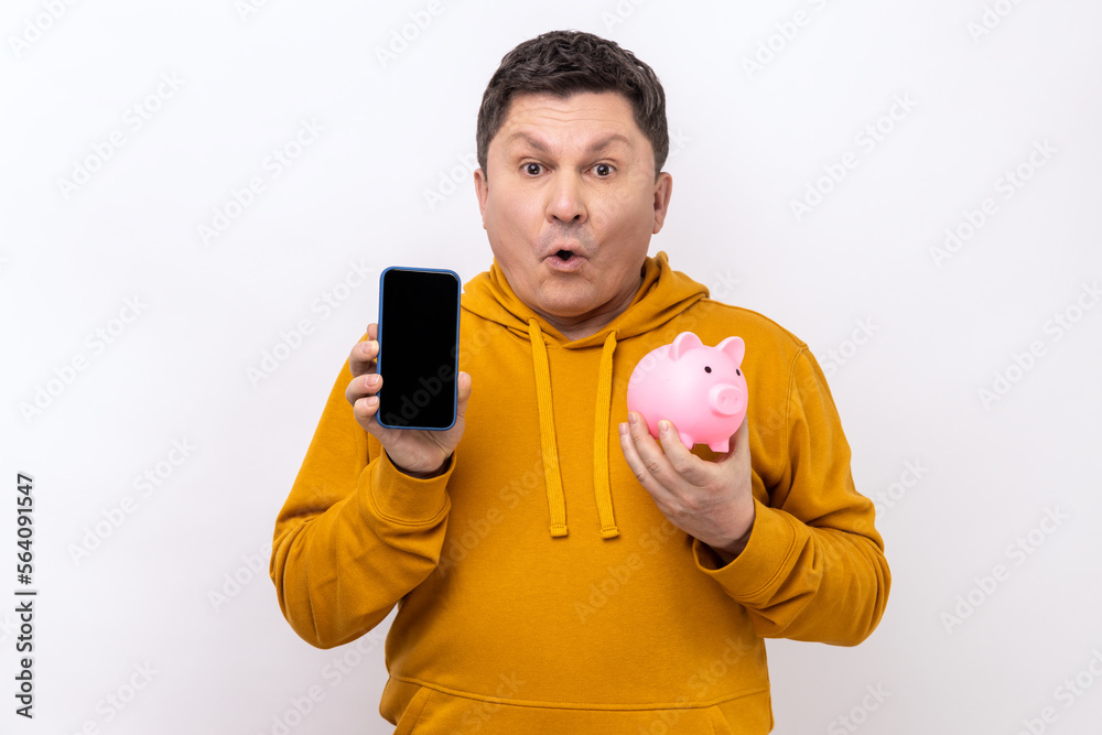 Astonished shocked man holding smartphone with empty screen and piggy bank, cashback, online wallet, wearing urban style hoodie. Indoor studio shot isolated on white background.