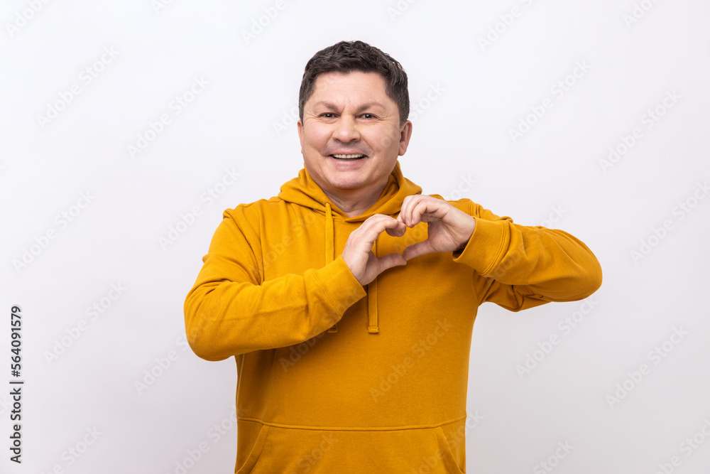 Happy handsome dark haired man standing with heart or love gesture and looking at camera with toothy smile, wearing urban style hoodie. Indoor studio shot isolated on white background. Happy handsome dark haired man standing with heart or love gesture and looking at camera with toothy smile, wearing urban style hoodie. Indoor studio shot isolated on white background.