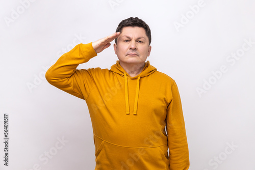 Subordinate man saluting, holding hand near head, looking at camera with serious expression, patriotism and discipline, wearing urban style hoodie. Indoor studio shot isolated on white background.