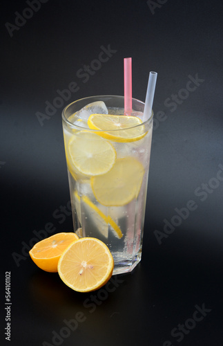 Lemonade, a refreshing drink in a tall glass with ice, straws and slices of ripe lemon on a black background.