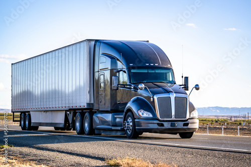 Black stylish big rig semi truck with truck driver sleeping compartment transporting cargo in dry van semi trailer driving on the flat highway road