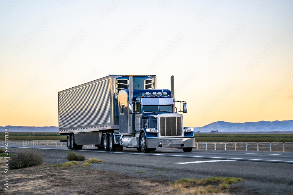 Heavy duty dark blue classic stylish big rig semi truck tractor with lot of chrome accessories transporting cargo in refrigerator semi trailer driving on the road at twilight - obrazy, fototapety, plakaty 