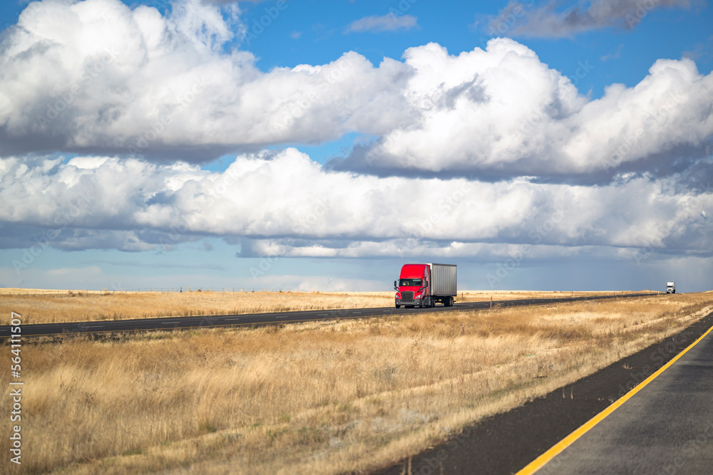 Industrial red big rig semi truck transporting loaded dry van semi trailer driving on the two branches road surrounded by yellowed fields Industrial red big rig semi truck transporting loaded dry van semi trailer driving on the two branches road surrounded by yellowed fields