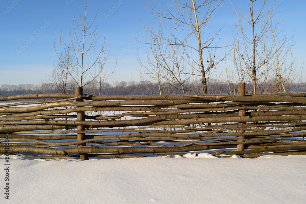 Fence made of small poles and linden branches on the background of a frozen river on a sunny day