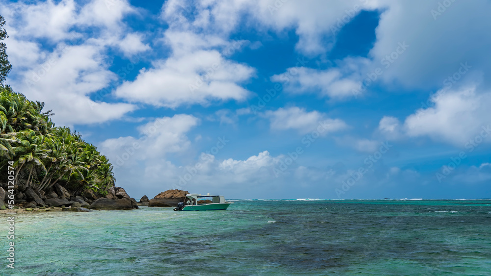 Fototapeta premium The boat is moored in the turquoise ocean off a tropical island. A scattering of boulders on the shore. On the slope- thickets of palm trees. Clouds in the blue sky. Seychelles. Moyenne Island