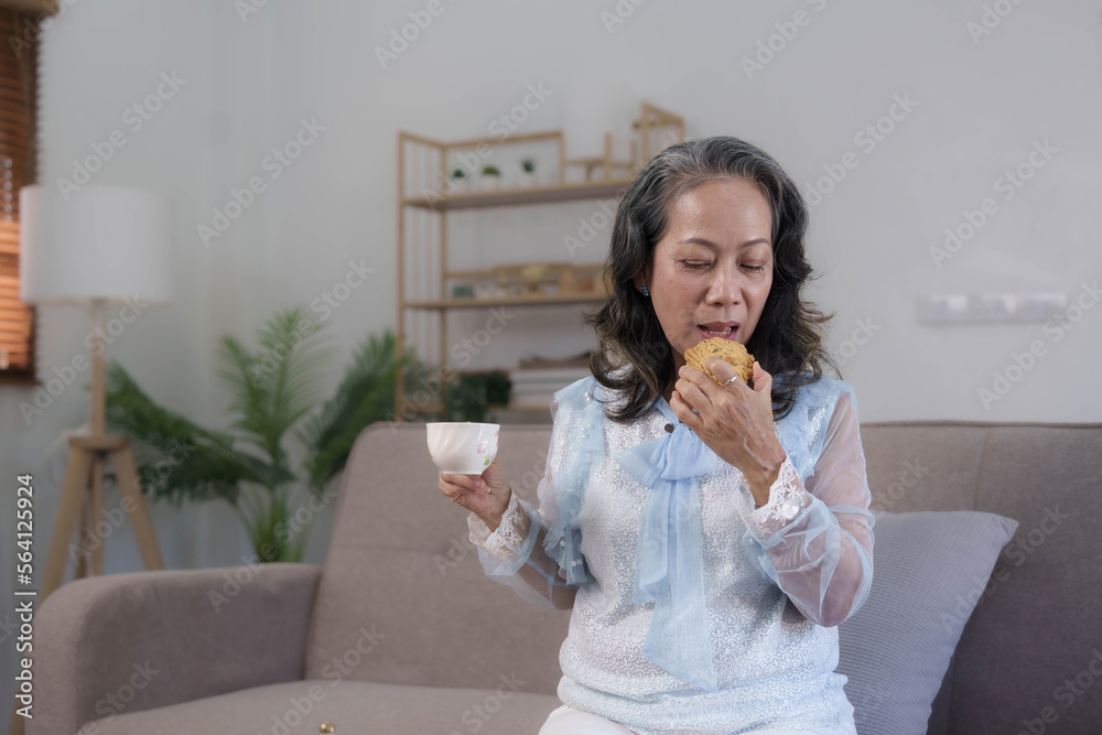 Beautiful and happy Asian 60s aged woman having breakfast, sipping a cup of tea and eating cookies in her living room. Beautiful and happy Asian 60s aged woman having breakfast, sipping a cup of tea and eating cookies in her living room.