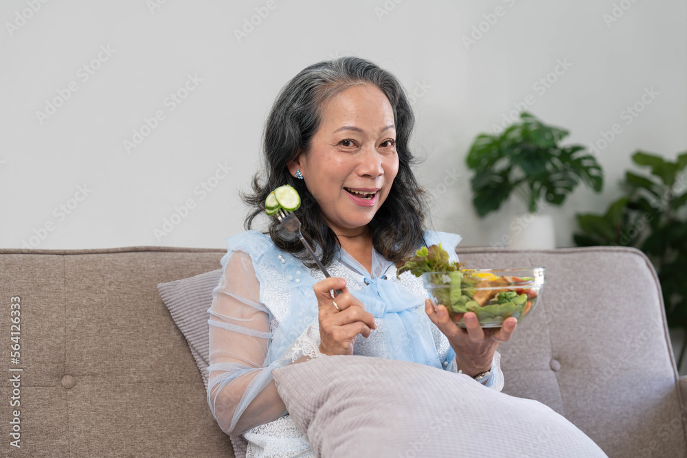 Portrait, Cheerful Asian 60s aged woman having breakfast in her living room, eating healthy salad vegetables mix bowl. Portrait, Cheerful Asian 60s aged woman having breakfast in her living room, eating healthy salad vegetables mix bowl.