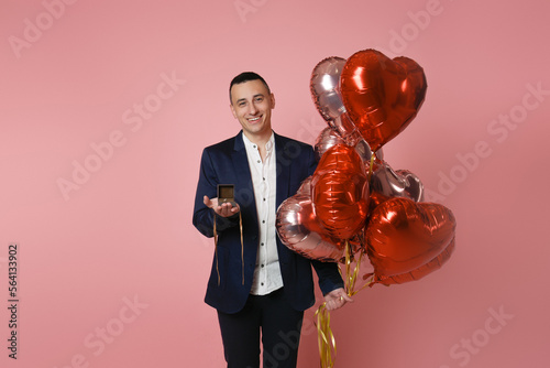 A man in a suit with red balloons on a pink background holds out a box with a wedding ring. Engaged on Valentine's Day, the man proposes, smiles. photo