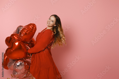 Beautiful girl in a red dress on a pink background, red and pink heart balloons. Joyful model having fun, celebrating, playing dances. Valentine's Day, February 14