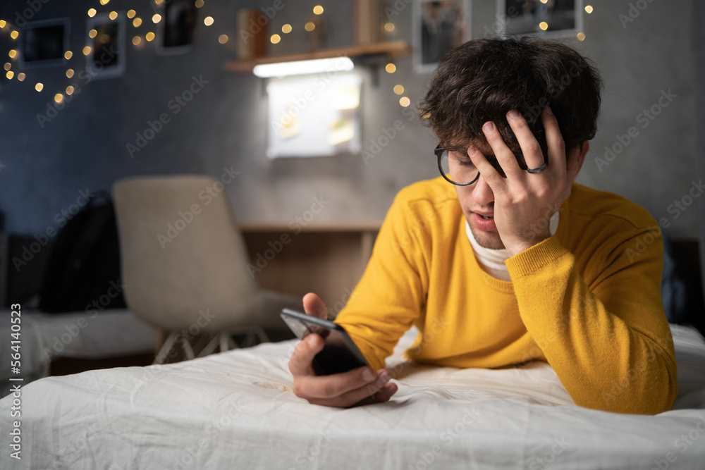 Young man reading bad news on phone while lying on bed in dormitory Young man reading bad news on phone while lying on bed in dormitory