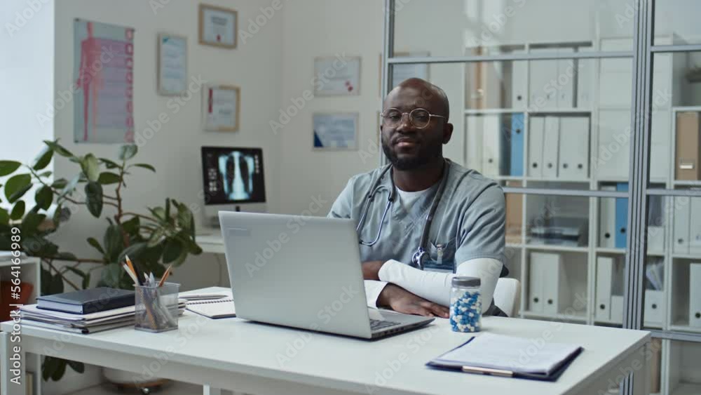 Young confident African American online doctor in uniform looking at camera while sitting by his workplace in front of laptop against shelves with medical documents and computer monitor on desk Young confident African American online doctor in uniform looking at camera while sitting by his workplace in front of laptop against shelves with medical documents and computer monitor on desk