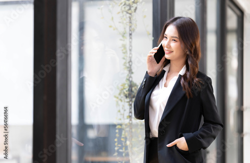 Happy young asian business woman wearing suit holding mobile phone standing in her workstation office