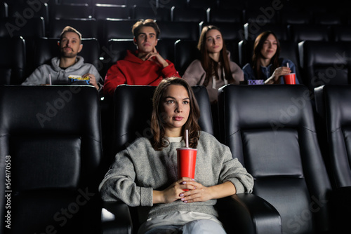 A young woman with a red cup in her hands attentively watches a movie. A woman came to the premiere at the cinema