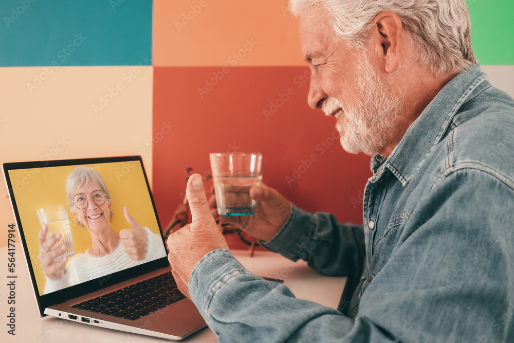 Smiling bearded old senior man chatting in online video using laptop holding a glass of water. Caucasian male having virtual video call with happy wife or friend. Colorful background, copy space Smiling bearded old senior man chatting in online video using laptop holding a glass of water. Caucasian male having virtual video call with happy wife or friend. Colorful background, copy space