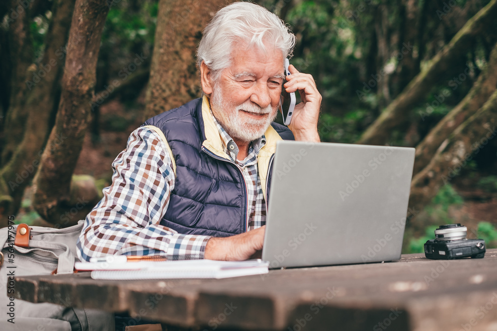 Smiling bearded senior man working on laptop using mobile phone while sitting at a wooden table outdoors in the middle of the park. Concept of smart working in nature, business in fresh air Smiling bearded senior man working on laptop using mobile phone while sitting at a wooden table outdoors in the middle of the park. Concept of smart working in nature, business in fresh air
