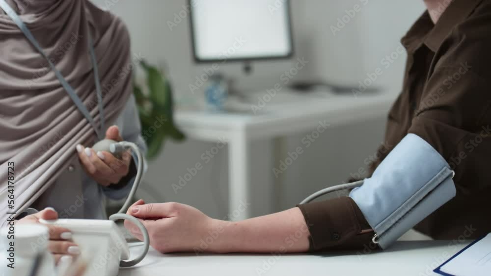 Close-up of young Muslim female physician or general practitioner in hijab checking blood pressure of patient while both sitting by workplace of clinician in medical office