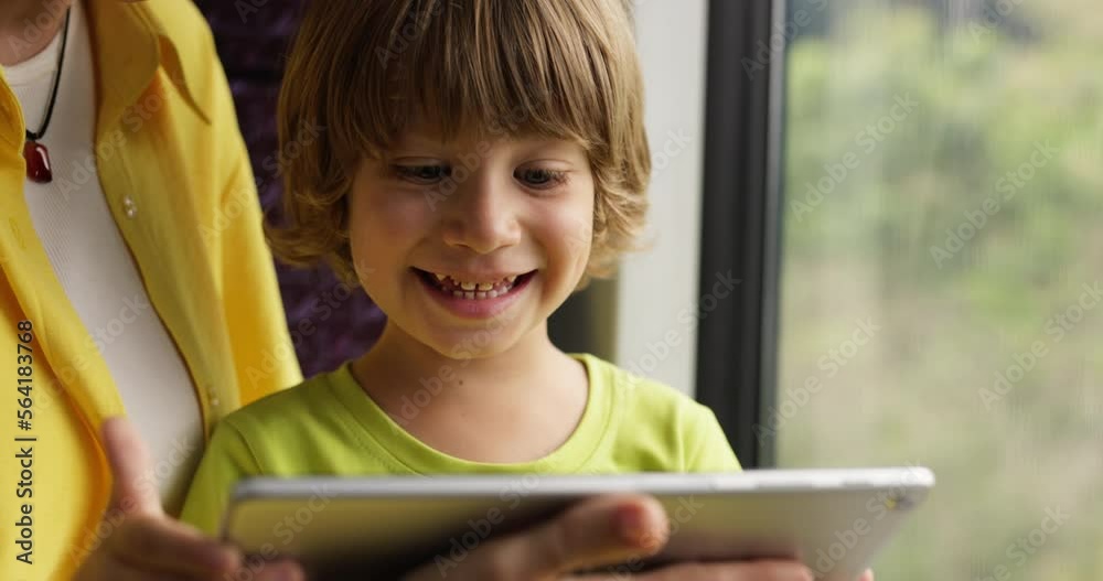 Child on bus goes on excursion and spends time watching videos on tablet with his mother. Boy is sitting at window in bus with mother and watching video on tablet, smiling, laughing and feeling great. Child on bus goes on excursion and spends time watching videos on tablet with his mother. Boy is sitting at window in bus with mother and watching video on tablet, smiling, laughing and feeling great.