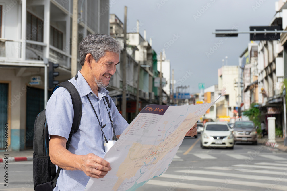elderly pensioner man with grey hair and beard carried backpack standing at the crossroads by the street,using city paper map to search tourist attractions,senior male enjoys travelling after retired