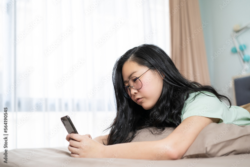 Beautiful glasses Asian woman lies on her stomach and plays mobile on the bed in her bedroom with a pastel green - brown color theme. Beautiful glasses Asian woman lies on her stomach and plays mobile on the bed in her bedroom with a pastel green - brown color theme.