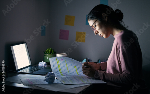 Student, reading notebook and studying at night with laptop of learning, education or internet project. Young woman, research paper and writing at dark desk for planning college homework in dormitory photo