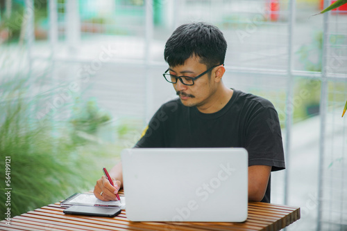 asian freelancer man wearing glasses, sits, working from home, writing and using a laptop and mobile phone in the home graden. this photo was taken on October 25, 2022 in yogyakarta city indonesia, 