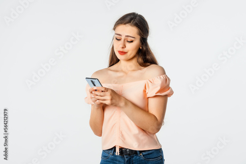 Smiling girl using mobile phone, holding telephone, looks happy, stands over white background. Pretty brunette female holding cell phone and reading funny message she received from boyfriend