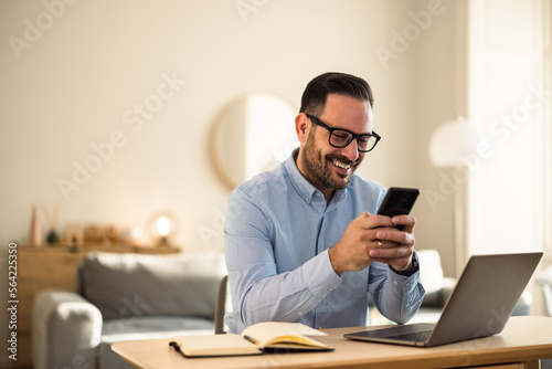 Smiling man typing on a mobile phone, sitting at the home office, with glasses on.