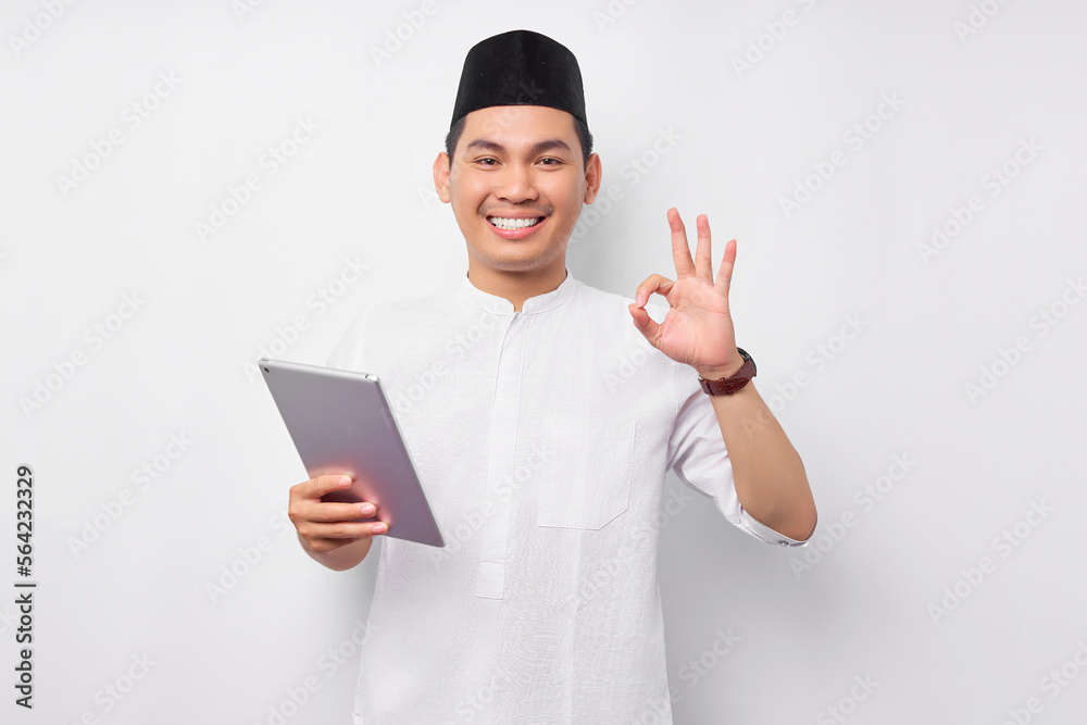 Smiling young Asian Muslim man holding digital tablet and showing okay sign isolated on white background. People religious Islam lifestyle concept. celebration Ramadan and ied Mubarak Smiling young Asian Muslim man holding digital tablet and showing okay sign isolated on white background. People religious Islam lifestyle concept. celebration Ramadan and ied Mubarak